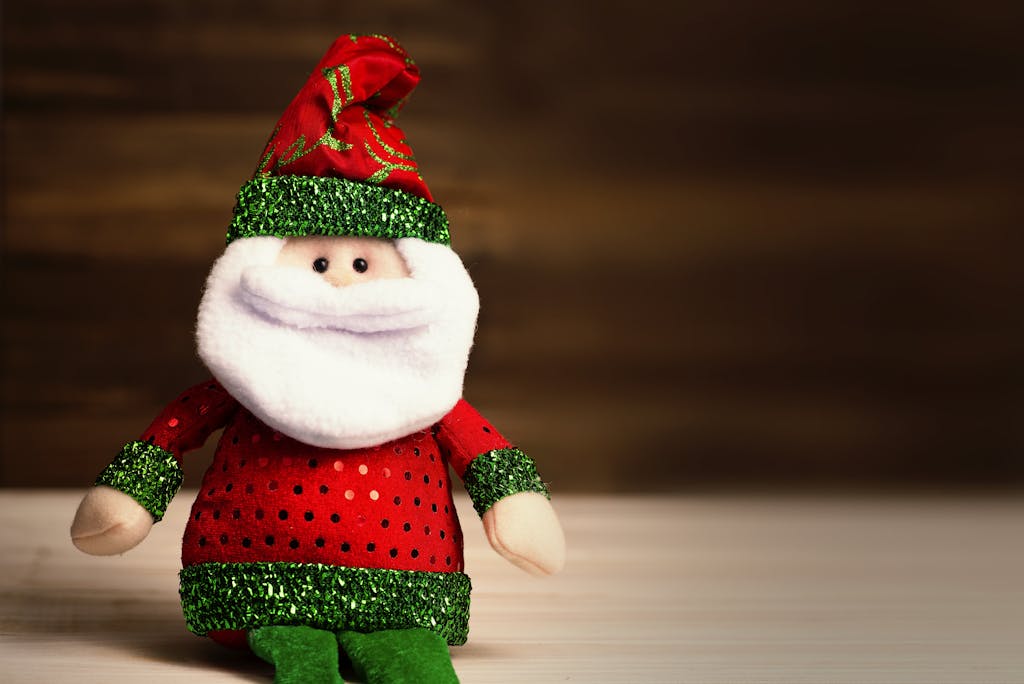 Cute Santa Claus plush toy wearing red and green attire sits on a wooden surface, perfect for holiday decor.