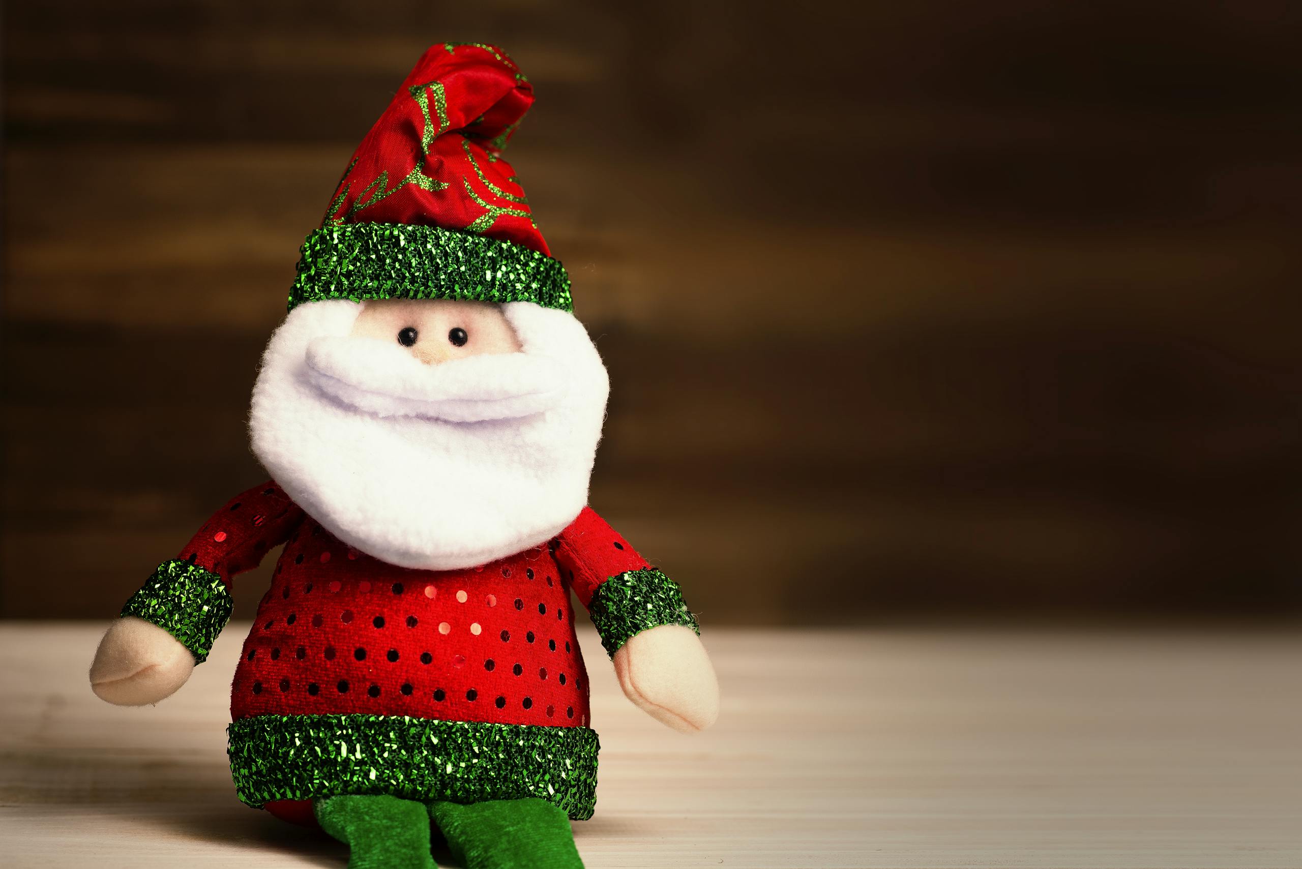 Cute Santa Claus plush toy wearing red and green attire sits on a wooden surface, perfect for holiday decor.